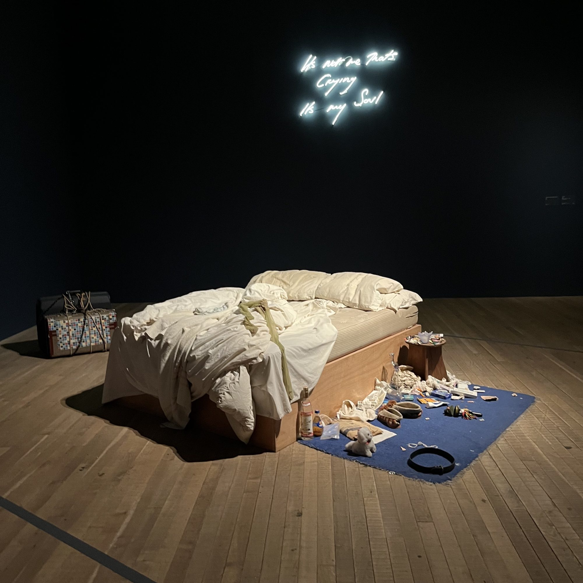 Installation view of My Bed by Tracey Emin. An unmade bed with tangled white sheets sits in a dark gallery space, surrounded by personal objects scattered across a blue rug, including bottles, tissues and clothing. Above the bed, a white neon text work glows against the black wall.

