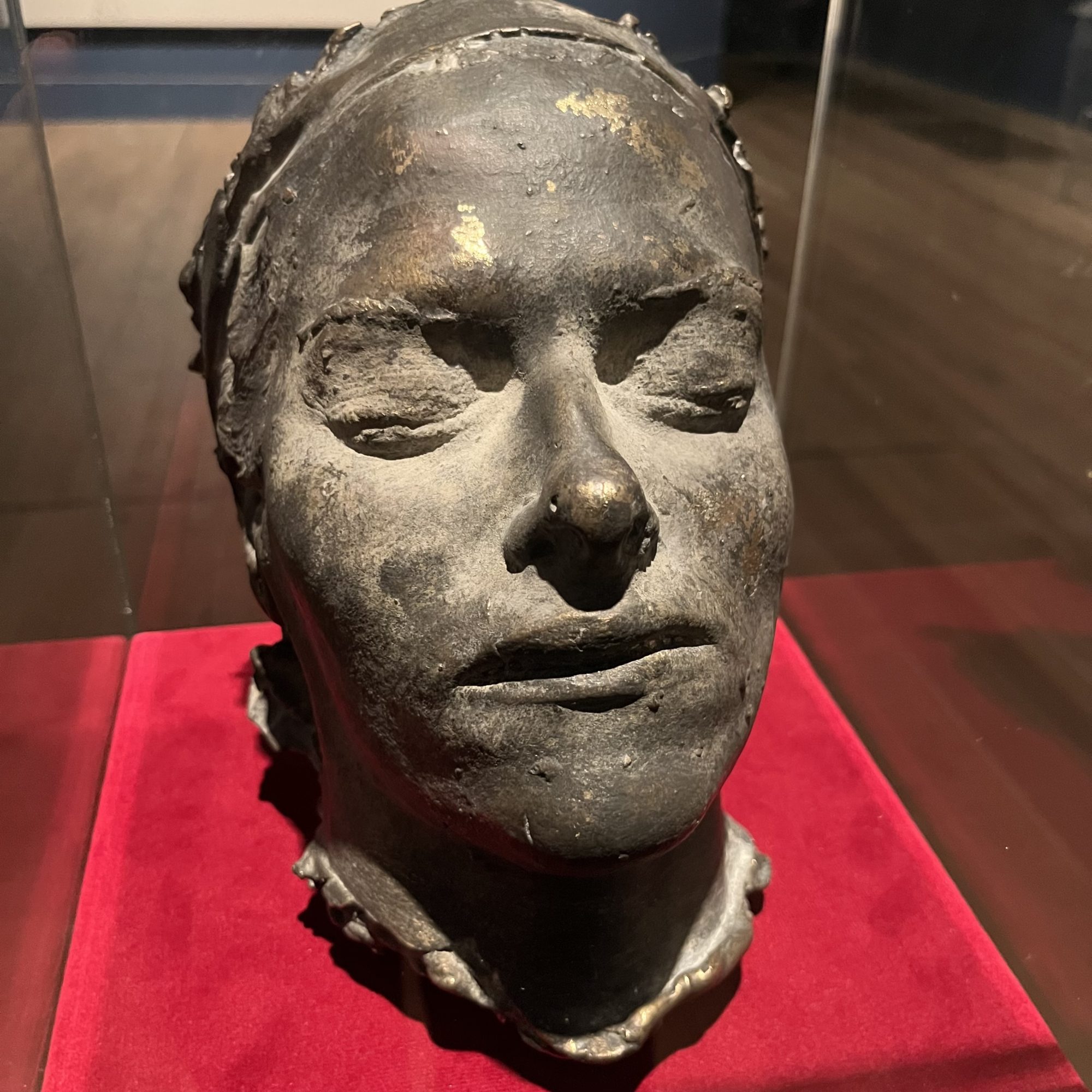 Close-up view of Death Mask by Tracey Emin. A dark bronze cast of a human face sits within a glass display case on a red velvet base. The surface appears rough and worn, with closed eyes and partially open lips creating a solemn, spectral expression.