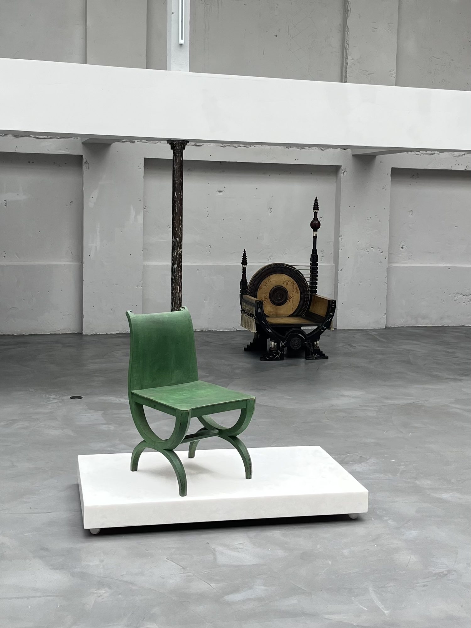 nstallation view showing a green Pierre Legrain chair displayed on a white platform, with Carlo Bugatti’s ornate Curule throne chair positioned behind it against a grey gallery wall.